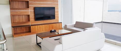 A modern living room with a flat-screen TV mounted on a wooden wall, a white sofa, and a glass coffee table.