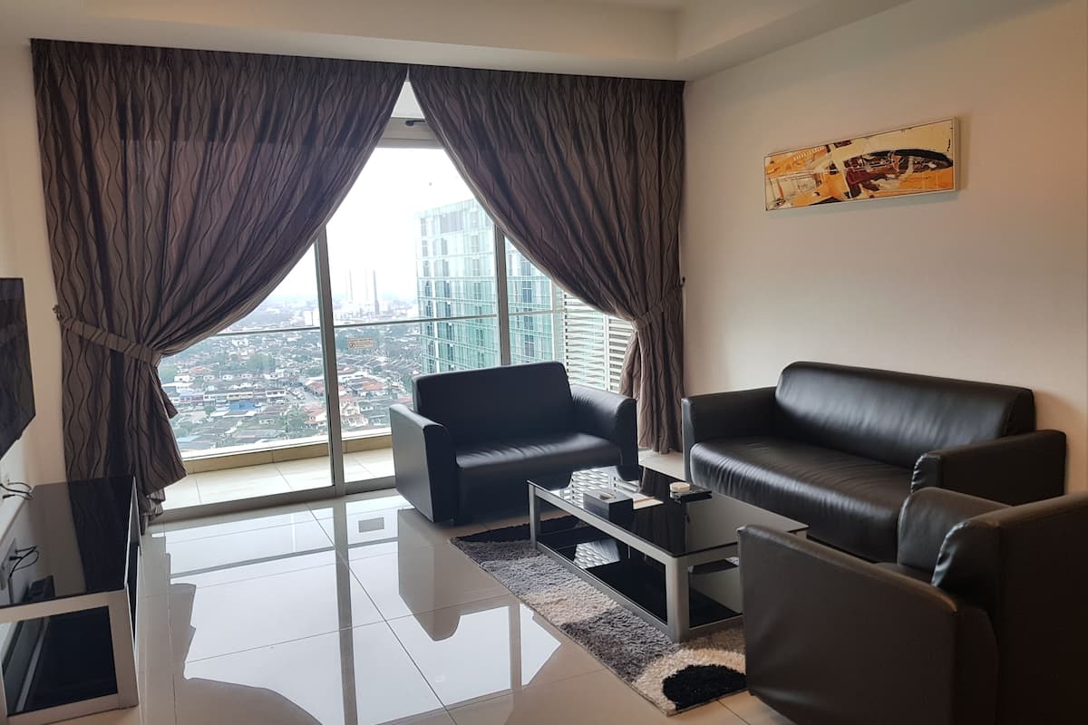 A modern living room with a black leather sofa, a black armchair, and a glass coffee table. Large windows with curtains offer a view of a cityscape.
