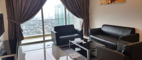 Una sala de estar moderna con un sofá de cuero negro, un sillón negro y una mesa de centro de vidrio. Amplias ventanas con cortinas ofrecen vistas a un paisaje urbano.