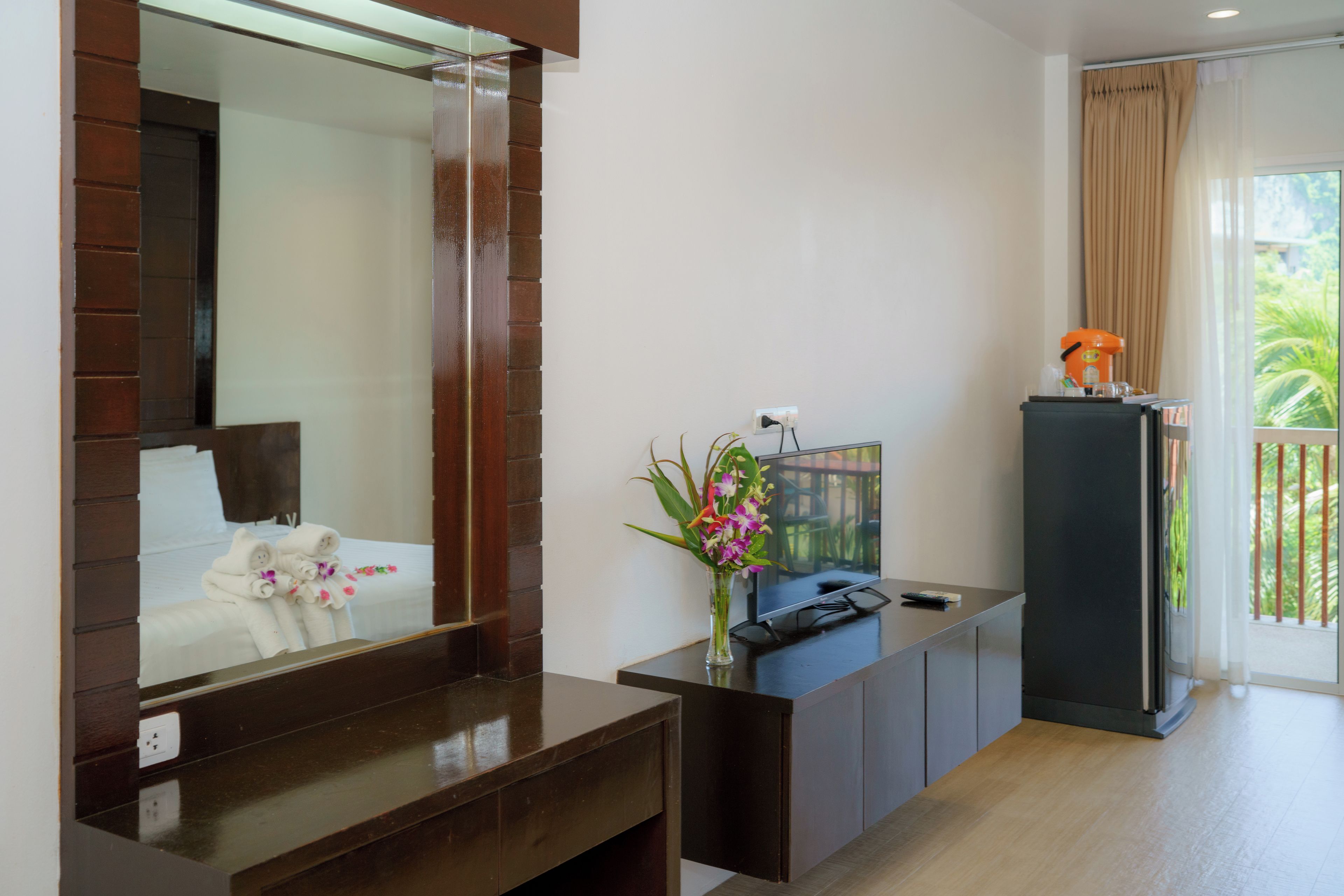A hotel room with a large mirror, a wooden vanity, a flat-screen TV, a small plant, and a balcony with curtains.