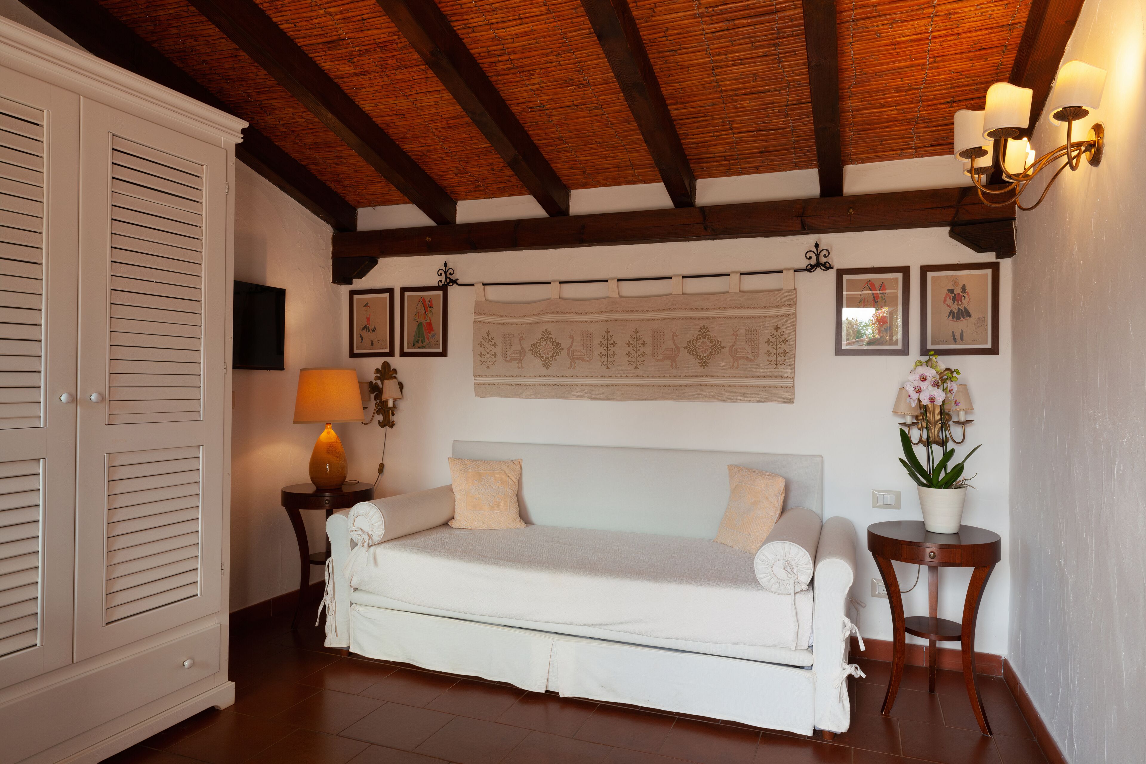 A cozy room with a wooden ceiling, a white sofa, a small table with a lamp, and a wall-mounted tapestry.