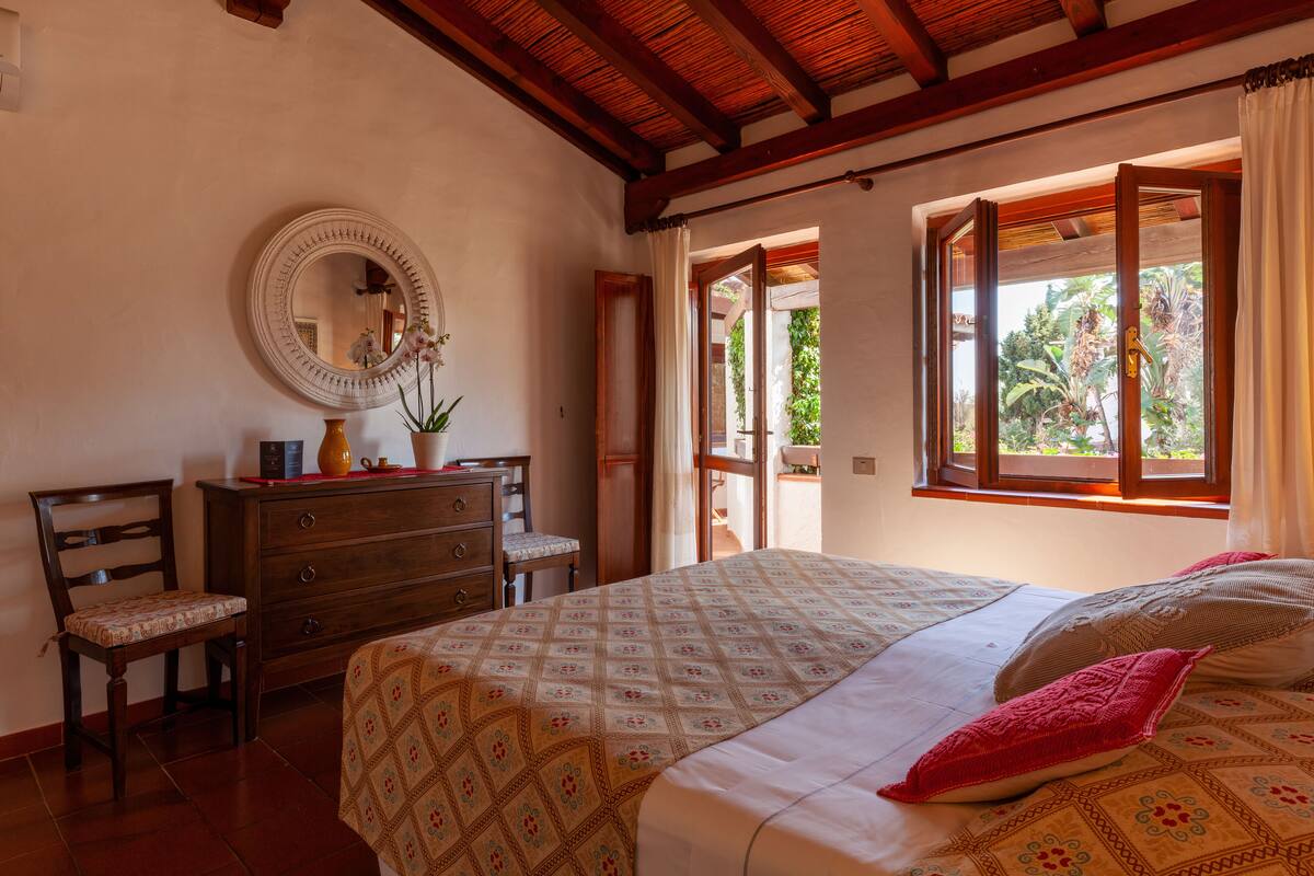 Une chambre à coucher avec un lit, une commode, une chaise, un miroir et une fenêtre avec des rideaux.