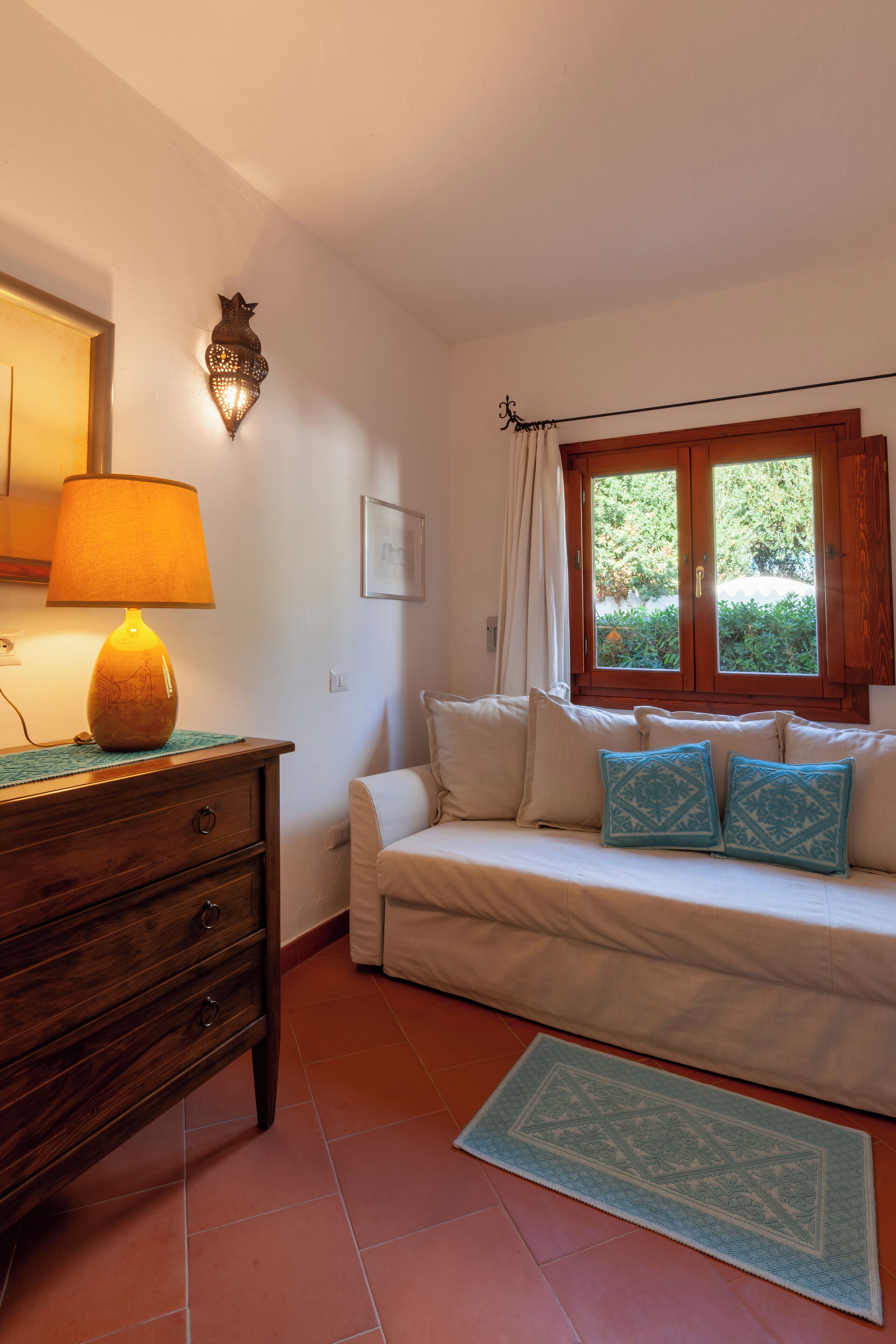 Una habitación con cómoda de madera, lámpara, sofá con cojines estampados, ventana con cortinas y una alfombra pequeña.