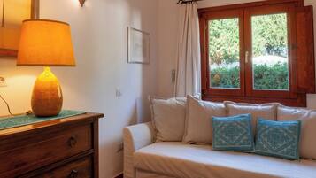 A room with a wooden dresser, a lamp, a sofa with patterned cushions, a window with curtains, and a small rug.