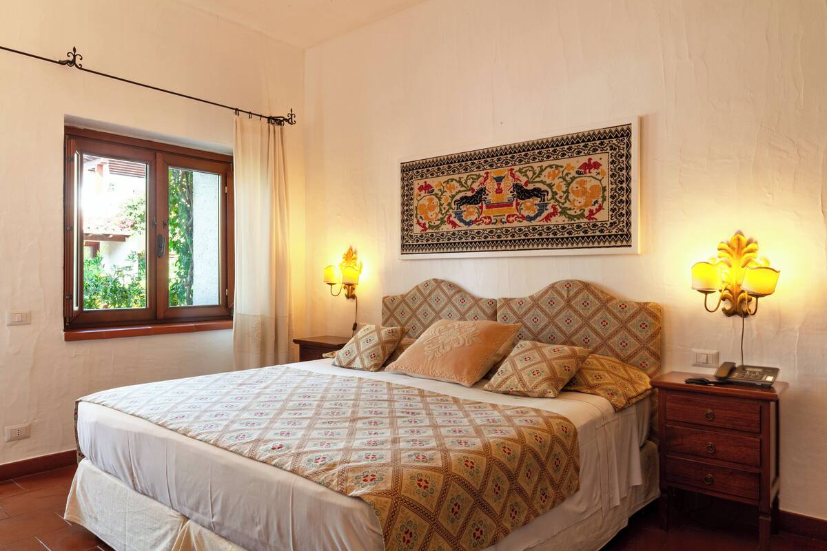 Une chambre à coucher comprenant un lit, deux tables de chevet, une lampe fixée au mur, une fenêtre avec des rideaux et un élément décoratif mural.