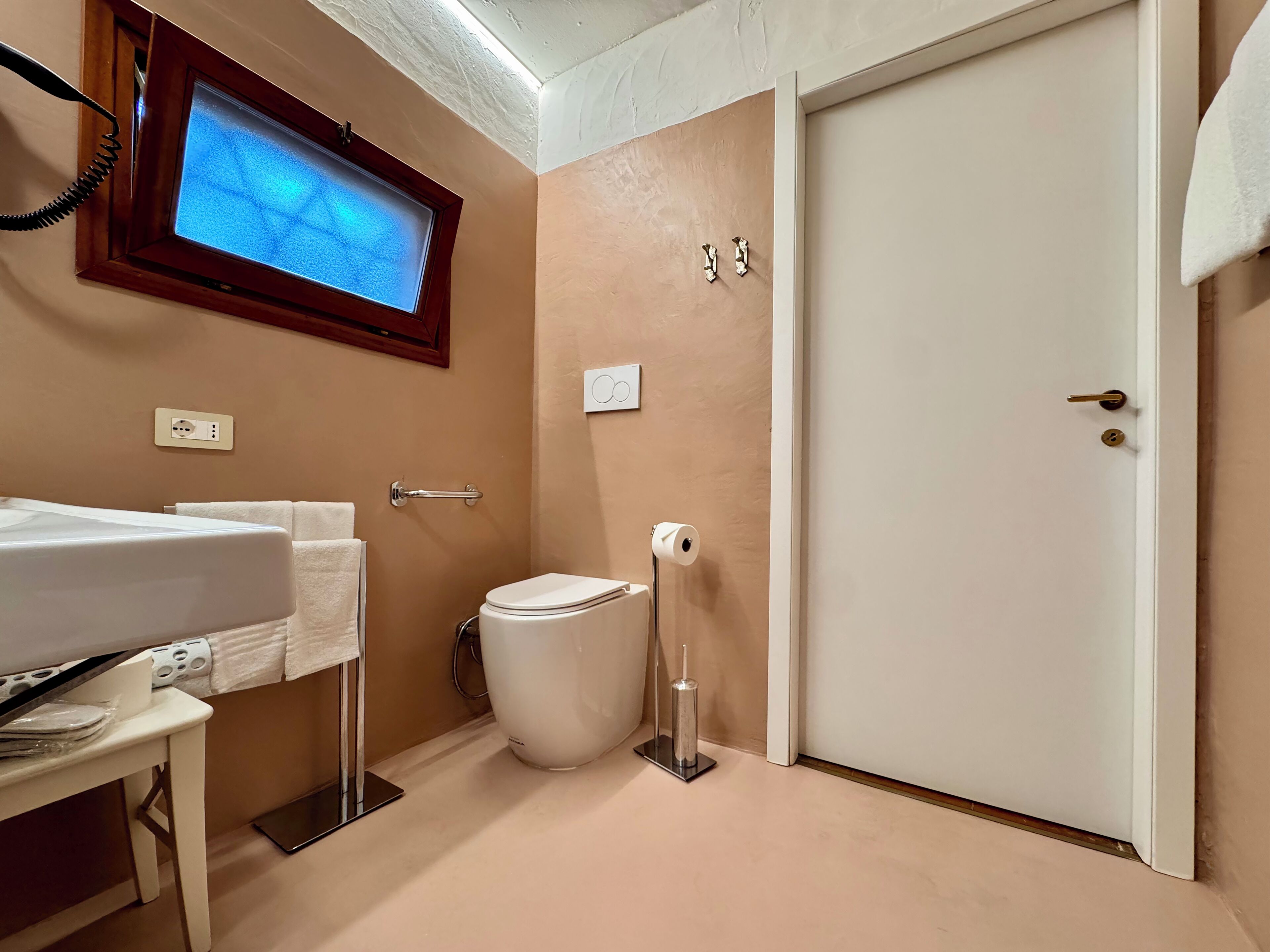 Un bagno con il water, un lavandino, uno specchio, un televisore montato sopra lo specchio e una porta con un asciugamano appeso.