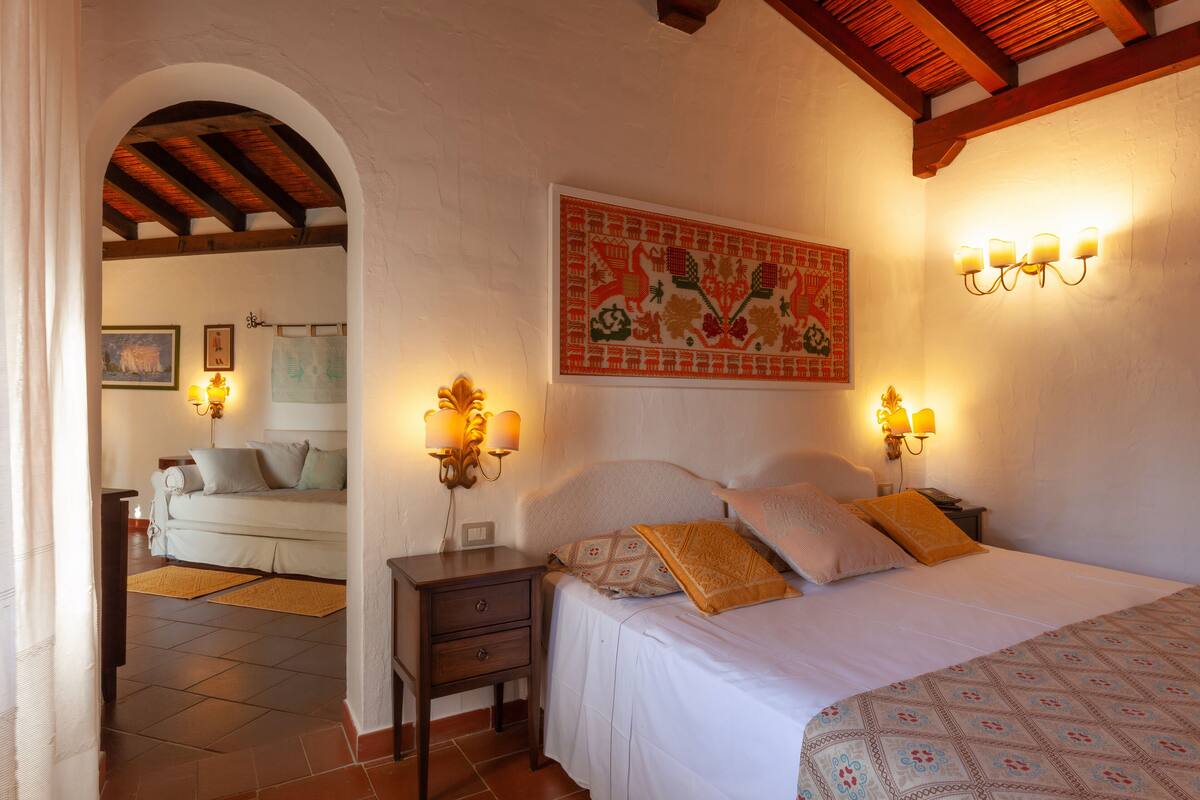 Une chambre à coucher avec un lit, des tables de chevet et des lampes fixées au mur. Il y a un tableau au mur et une porte qui donne sur une autre pièce.