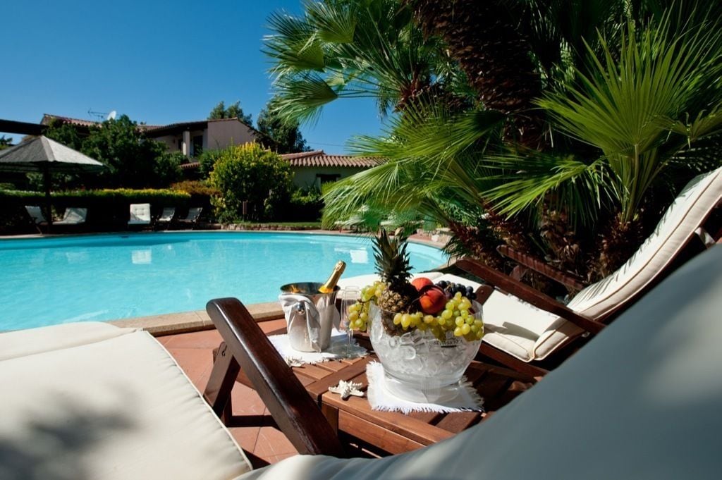 A poolside lounge area with a view of a swimming pool, surrounded by palm trees and a building in the background.