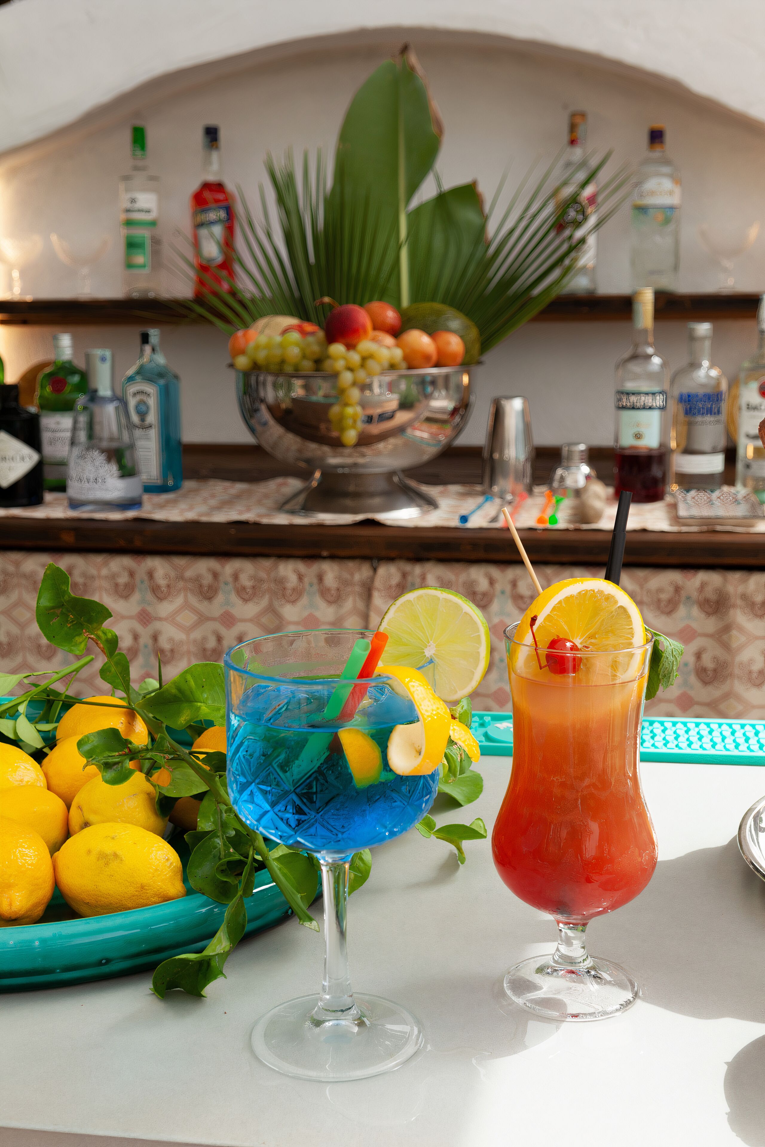 Two cocktails on a table with a fruit bowl and bottles in the background.