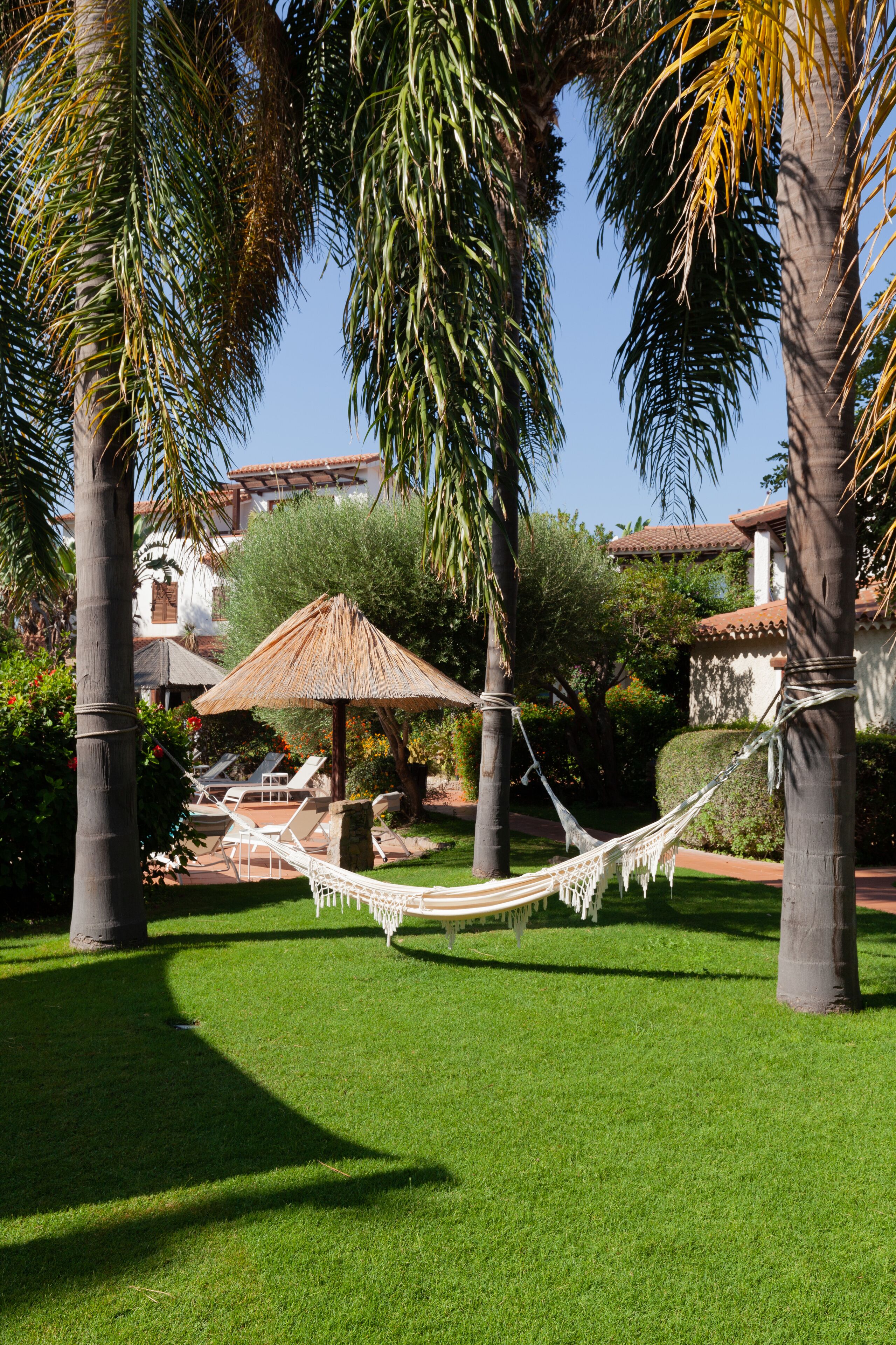Una hamaca entre palmeras, un toldo de paja y una zona de piscina con sillones de descanso.