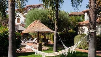 Una hamaca entre palmeras, un toldo de paja y una zona de piscina con sillones de descanso.