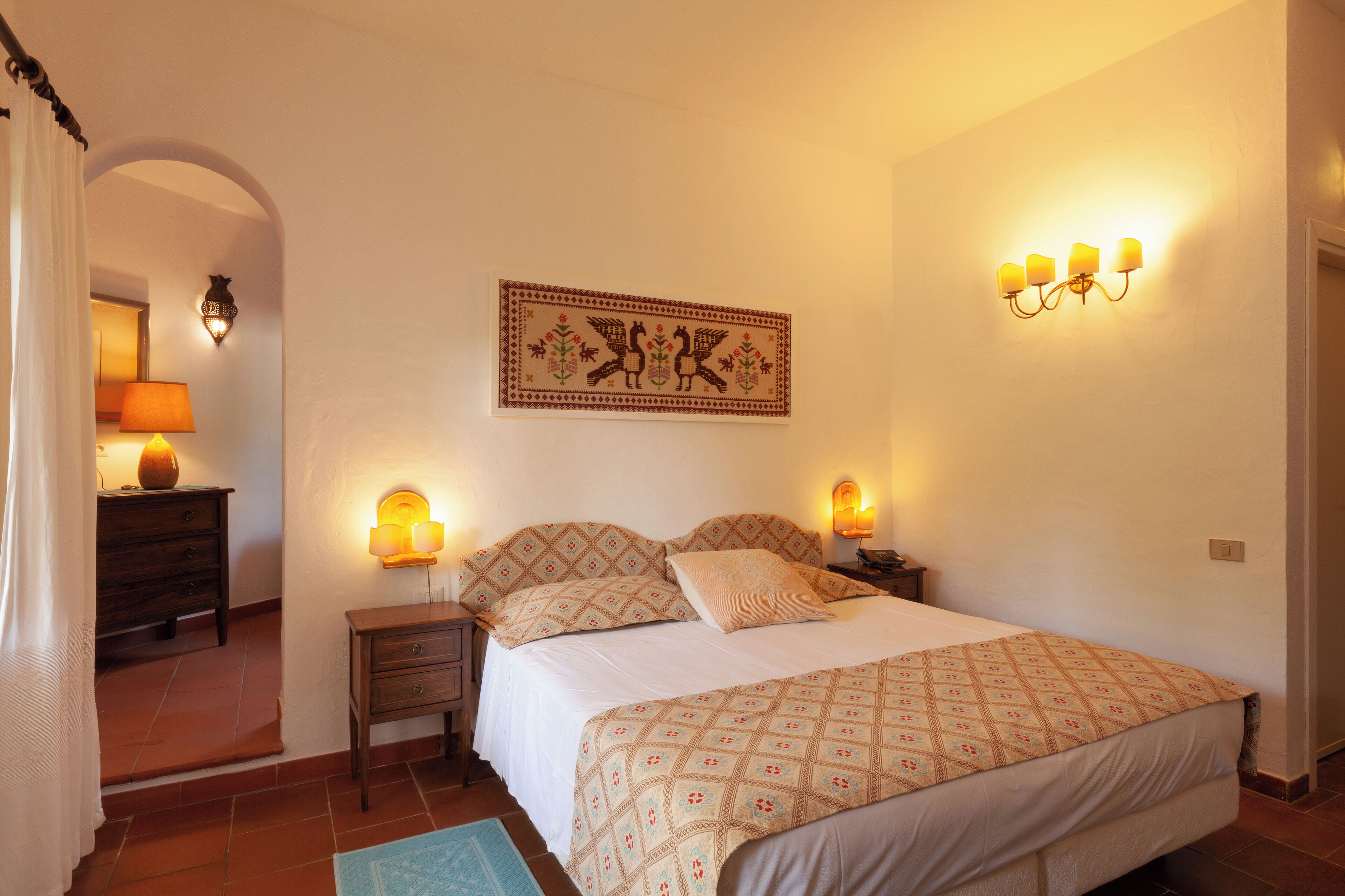 A bedroom with a bed, nightstands, a wall-mounted lamp, and a decorative tapestry.