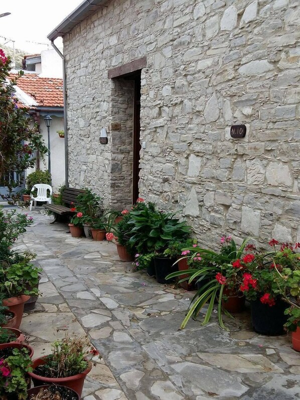 A stone building with a narrow stone-paved courtyard. There are potted plants and a white chair outside.