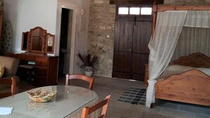 A rustic room with a stone wall, a wooden bed with a canopy, a round glass table with chairs, and a wooden cabinet.