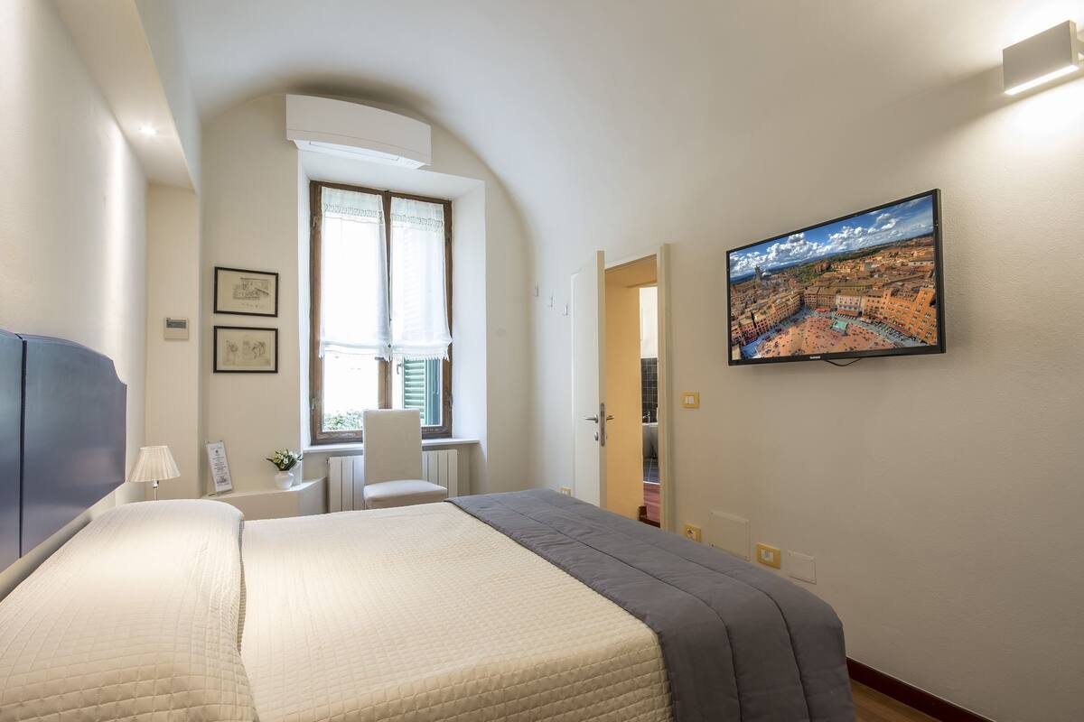 A hotel room with a bed, a desk, a chair, a window, and a TV on the wall.