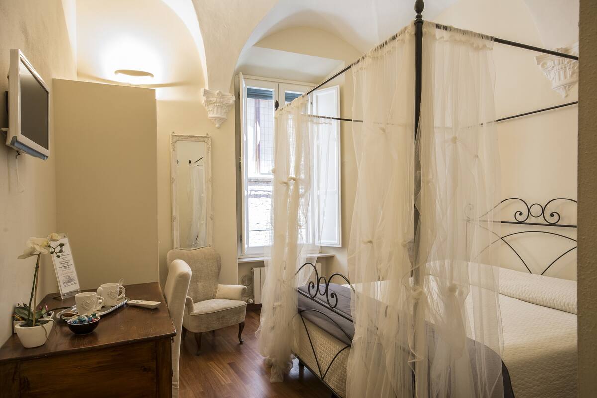 A bedroom with a bed, a chair, a desk with a computer, and a window with curtains.