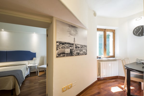 A room with a bed, a chair, a table, a clock, and a window with shutters.