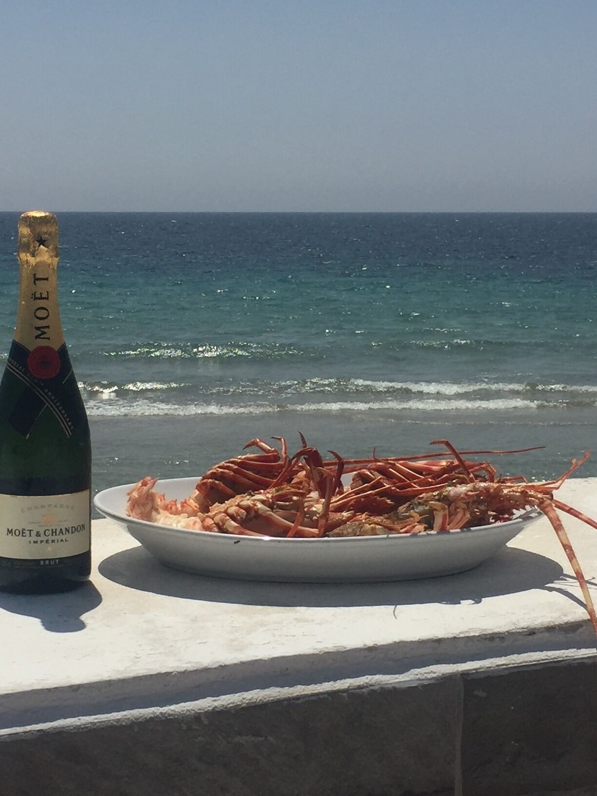 Une bouteille de champagne Moët & Chandon Impérial et un plat de homard cuit, le tout sur un balcon avec vue sur l’océan.