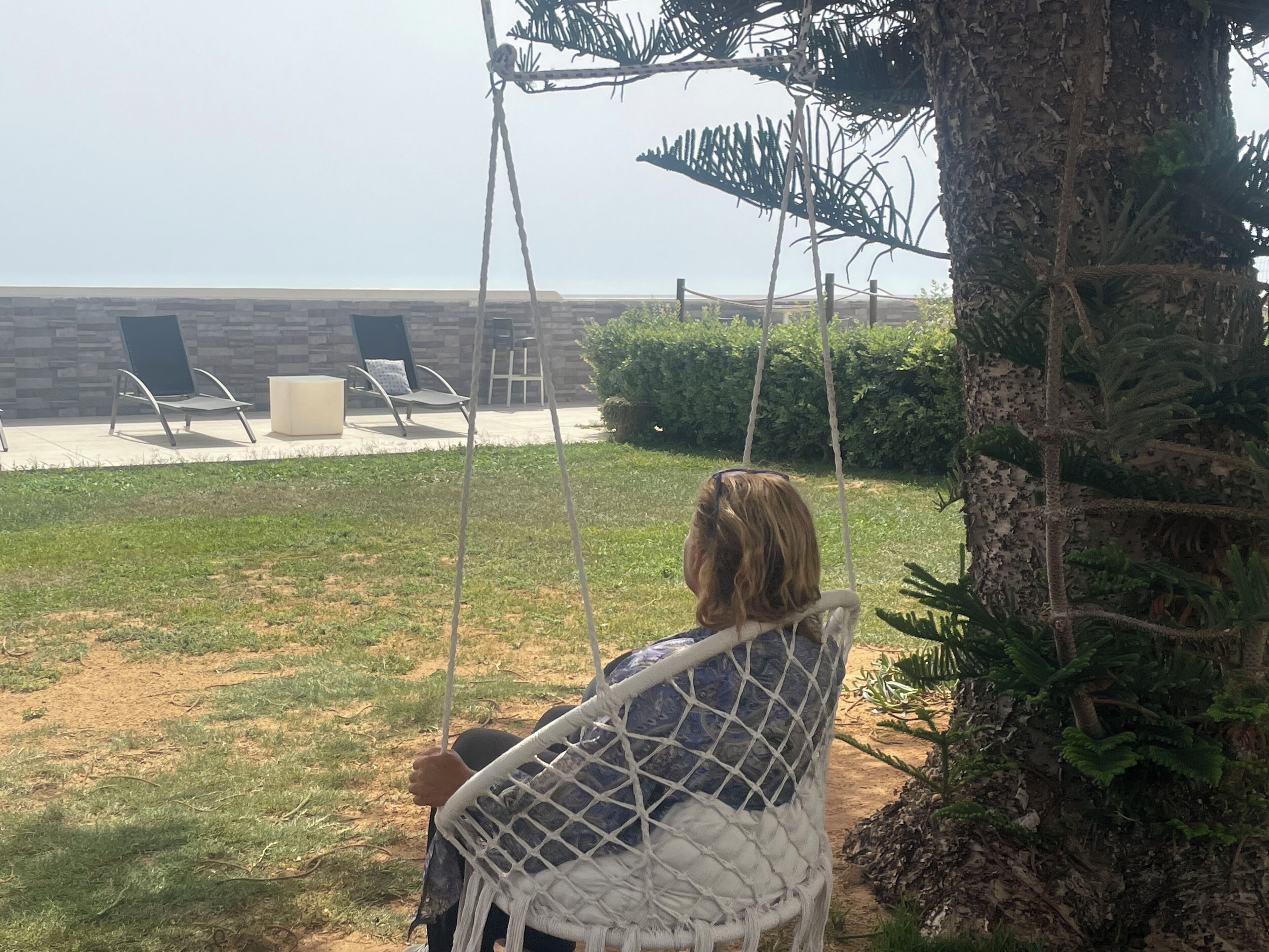 A hammock swing attached to a tree in a backyard with a view of a brick building and outdoor seating area.