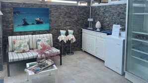 A modern room with a stone wall, a sofa, a coffee table with books, a painting of a beach, a minibar, and a glass door refrigerator.