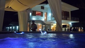 A pool area with a person standing near the edge, a building with balconies, and a group of people gathered outside.