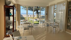 A dining area with a view of the sea, a bookshelf, and a table with chairs.