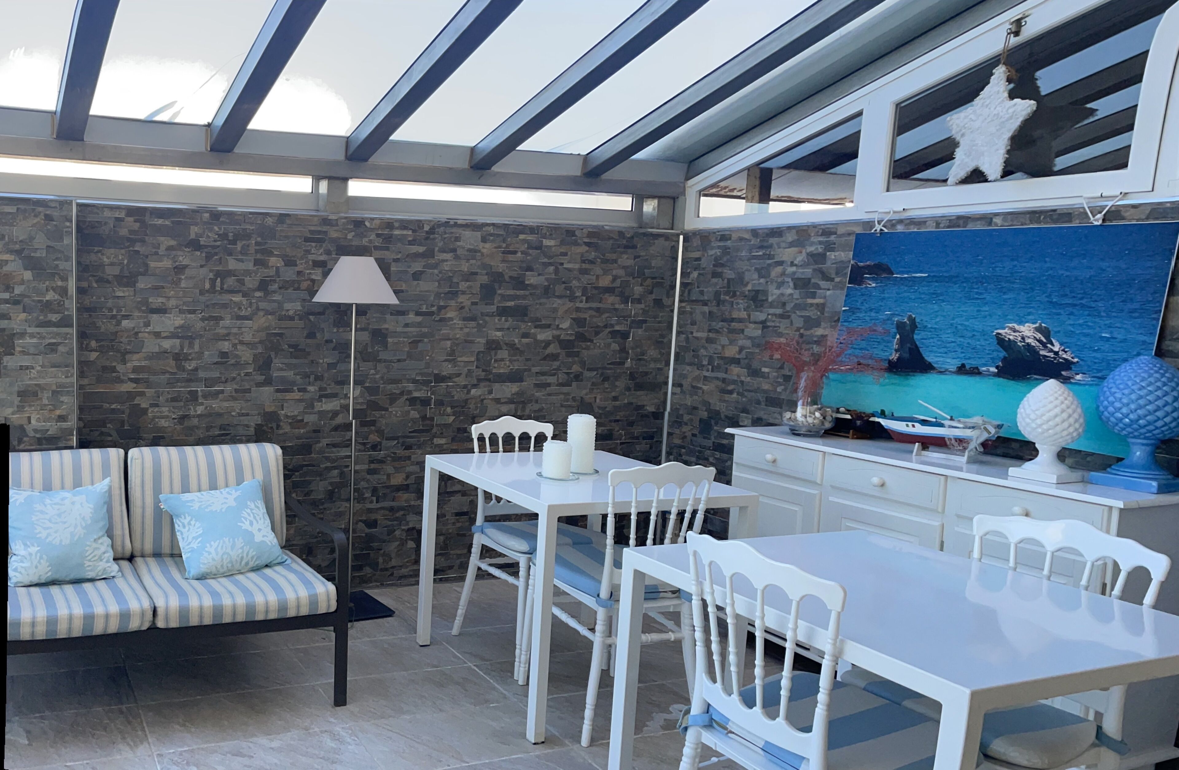 Un patio luminoso con un muro in pietra, un divano a righe, un tavolo da pranzo e un quadro raffigurante una scena marina.