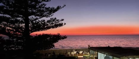 A beachfront property with a patio area, a large window overlooking the sea, and a tree in the foreground.