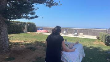 Une personne s’occupe d’un enfant assis sur une chaise longue, enveloppé dans une couverture, et ils sont tous les deux face à une plage et à d’autres chaises.