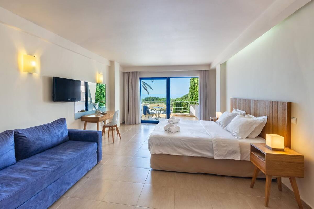 A hotel room with a bed, a sofa, a TV, a desk, and a balcony with a view.