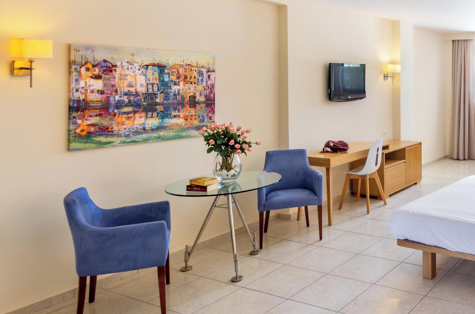 Une chambre d’hôtel comprenant un lit, un bureau, deux chaises, une table en verre avec un vase de fleurs, un tableau accroché au mur et un téléviseur fixé au mur.