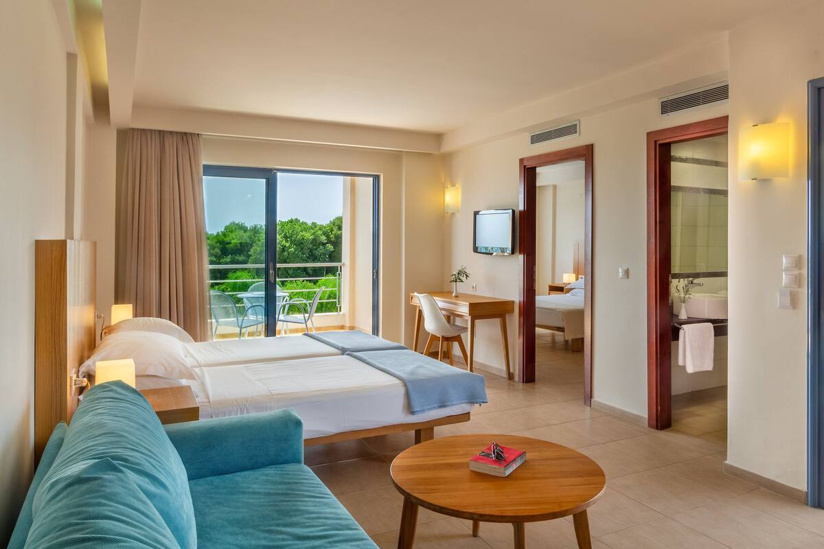 A hotel room with a bed, a desk, a chair, a small table, a TV, and a balcony with a view of greenery.