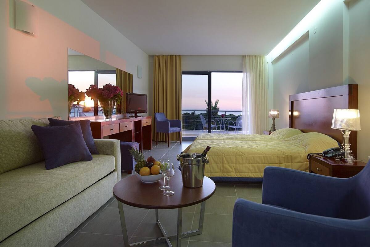 A hotel room with a bed, a sofa, a desk, a table with fruit and a drink, and a view of the sunset.