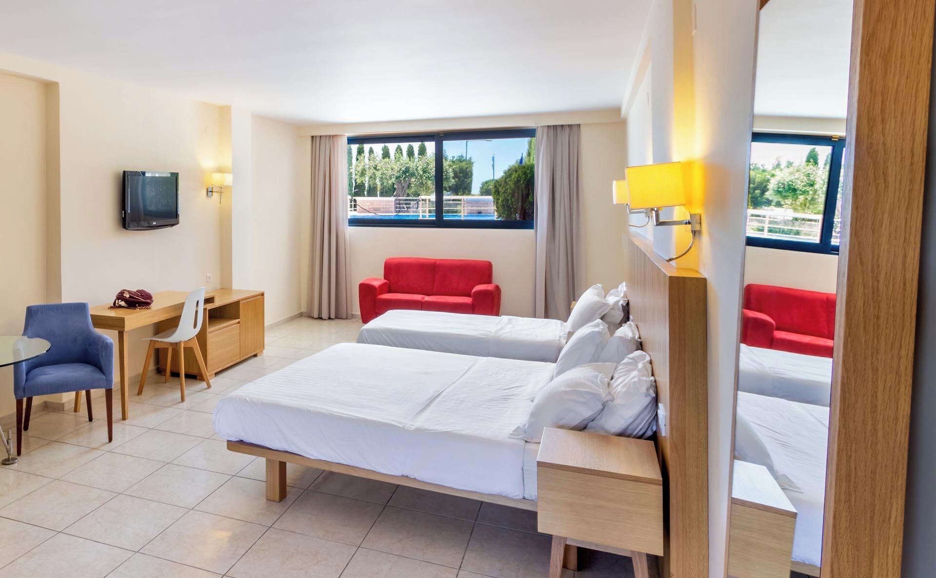 A hotel room with two beds, a desk, a chair, a TV, and large windows with curtains.