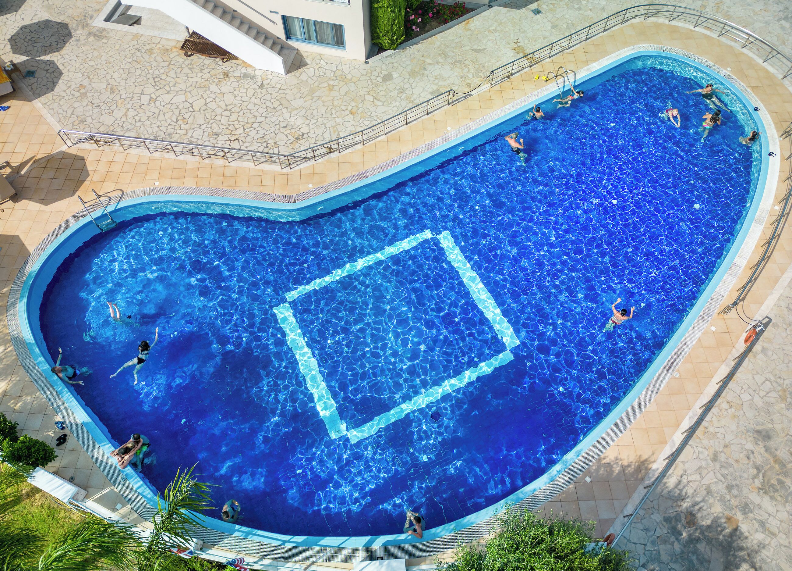 An aerial view of a heart-shaped swimming pool with clear blue water, surrounded by a stone deck and a building with a white facade.