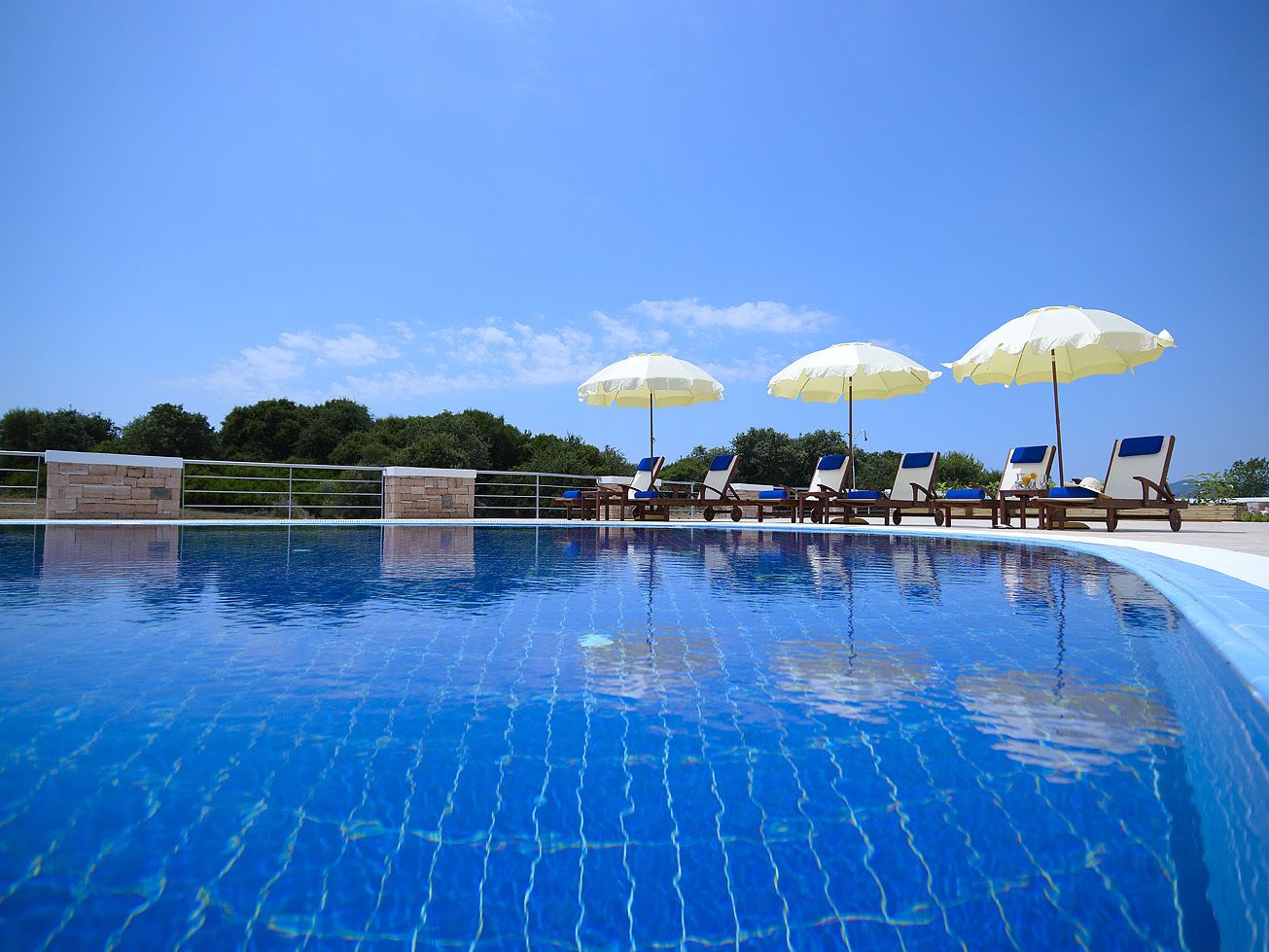 A swimming pool with clear blue water, surrounded by lounge chairs and umbrellas, and a stone wall in the background.