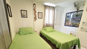 A room with two single beds, each with a green bedspread. There are framed pictures on the wall, a curtain, and a fish tank on the right side. - Posada Nativa Lizard House (San Andrés)