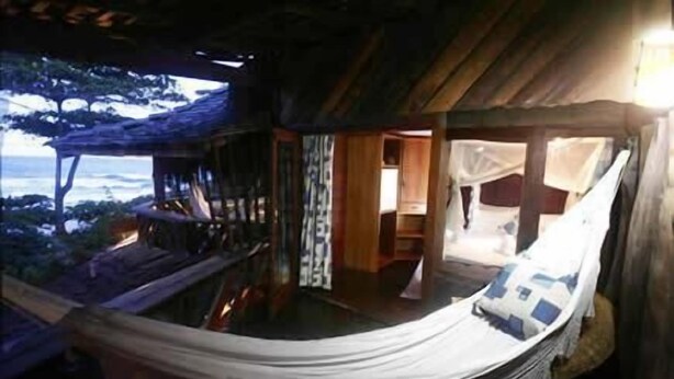 Una hamaca con vistas al océano, paredes de madera y una habitación pequeña con una cama y una mesita de noche.