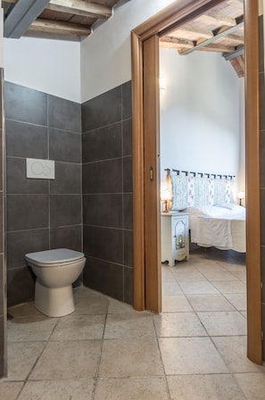 A bathroom with a toilet and a shower area, a bedside table with a lamp, and a bed with a patterned headboard. - I Terzi di Siena (Siena)