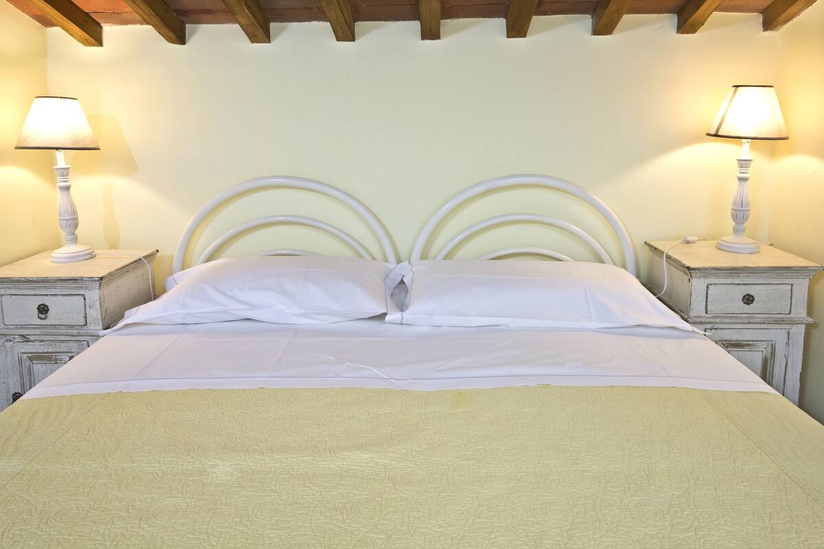 Uma cama bem arrumada com lençóis brancos e colcha amarela, acompanhada por duas mesas de cabeceira com abajures, em um quarto com design simples e clean.