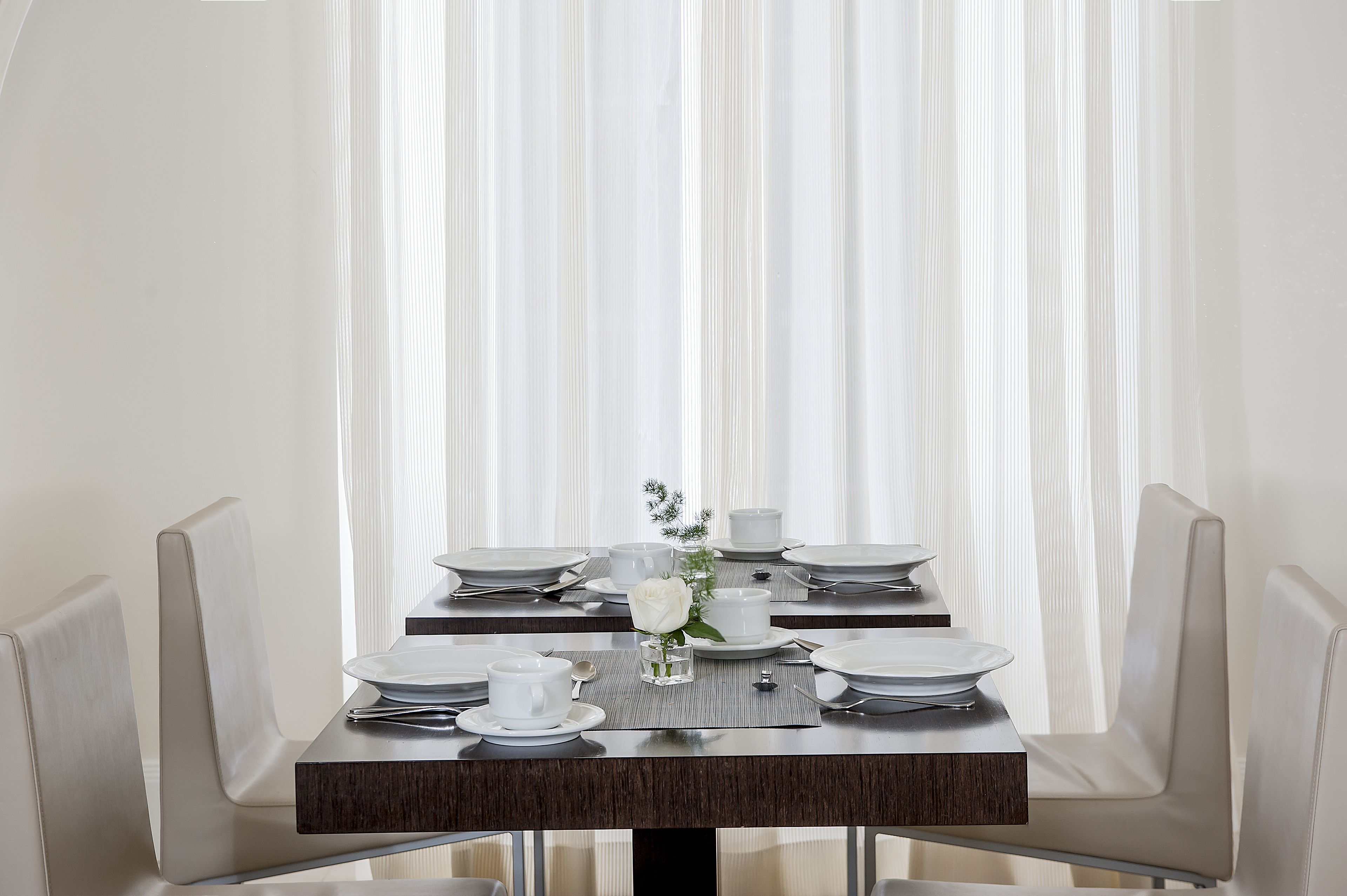 A dining table set for four with white plates, cups, and cutlery, a vase with a single white flower, and a small glass of water.