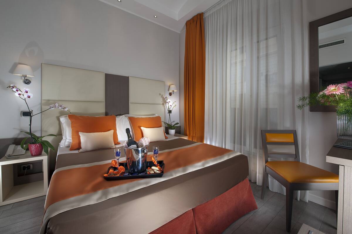 A modern hotel room with a large bed, a desk, and a chair. There is a tray with drinks and snacks on the bed.