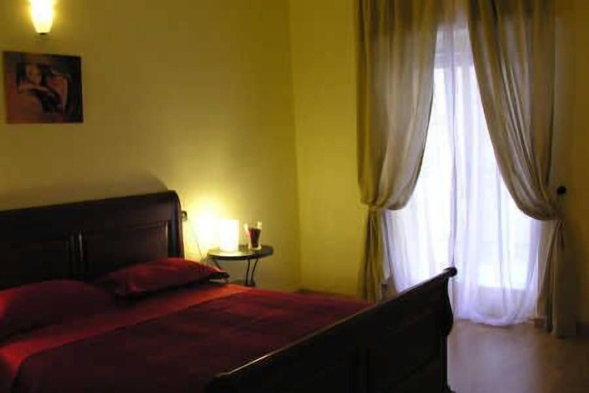 Una stanza con un letto, un comodino con una lampada e una finestra con tende.