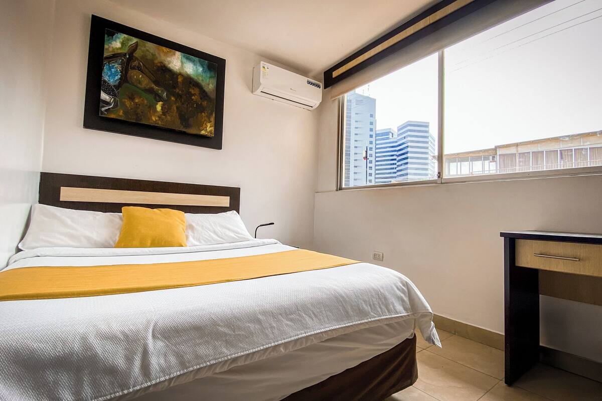 Habitación de hotel con cama, escritorio, ventana con vista a la ciudad y equipo de aire acondicionado.