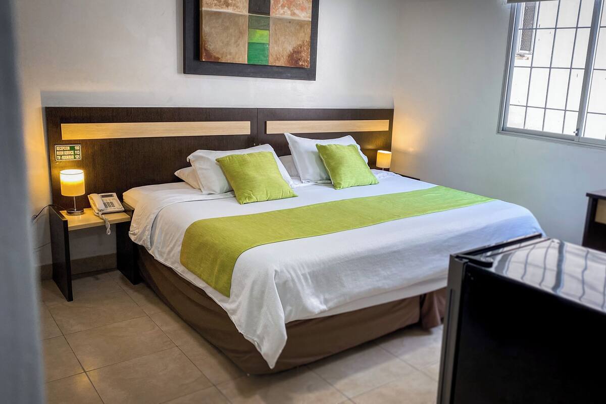 Una habitación de hotel con una cama, una mesita de noche con lámpara, un escritorio y una ventana con barrotes.