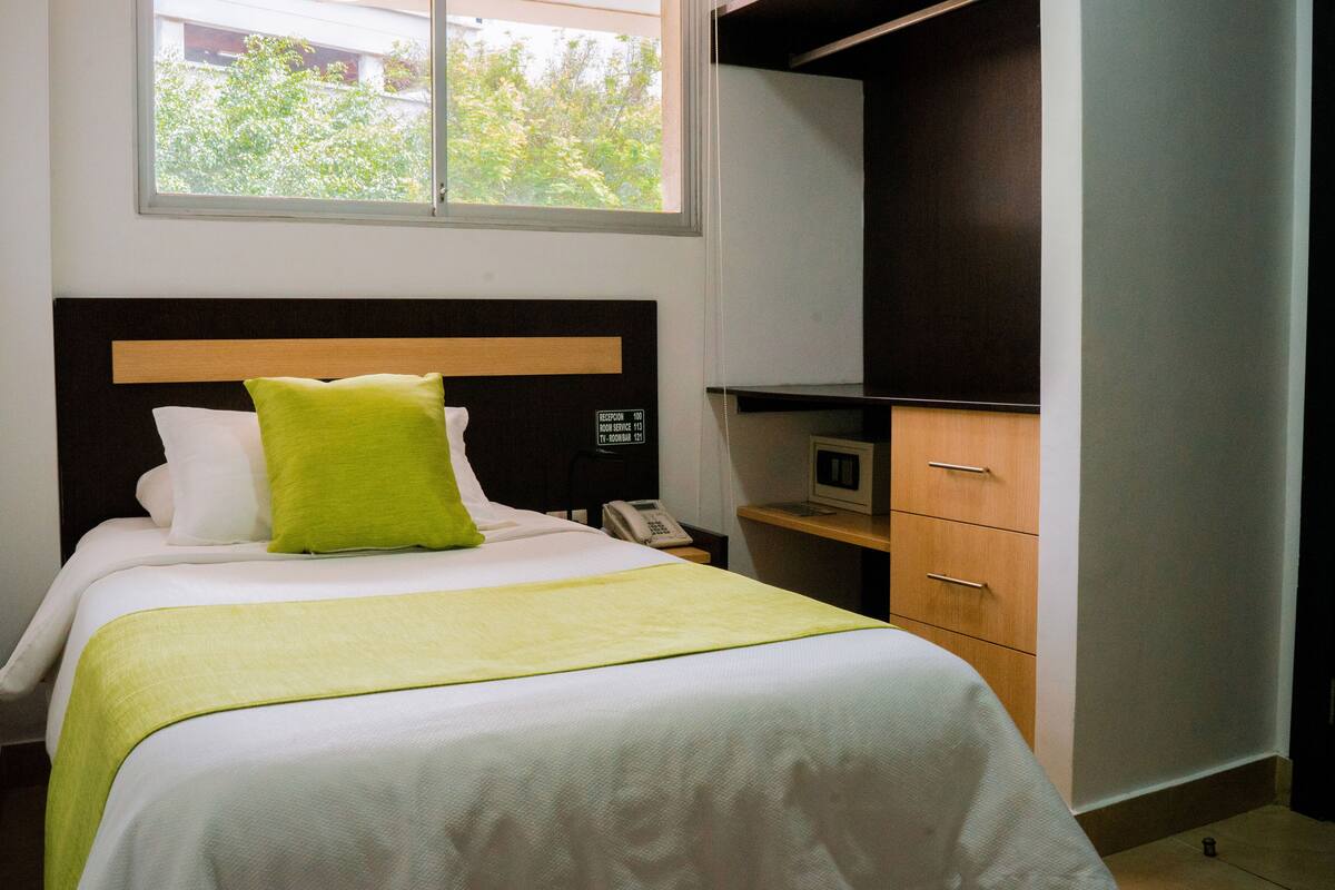 Una habitación de hotel con una cama, una mesita de noche con teléfono, un escritorio pequeño con computadora y una ventana con vistas a árboles.