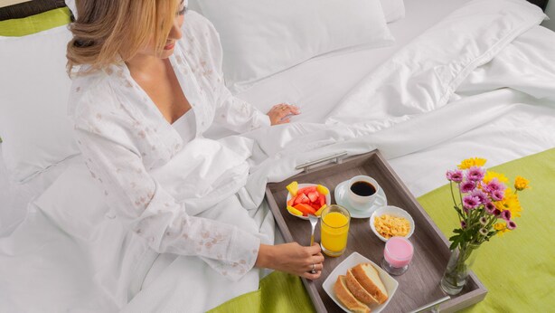 Eine Person in einem weißen Bademantel sitzt auf einem Bett mit einem Tablett mit Frühstücksgegenständen wie Obst, Saft und Müsli.