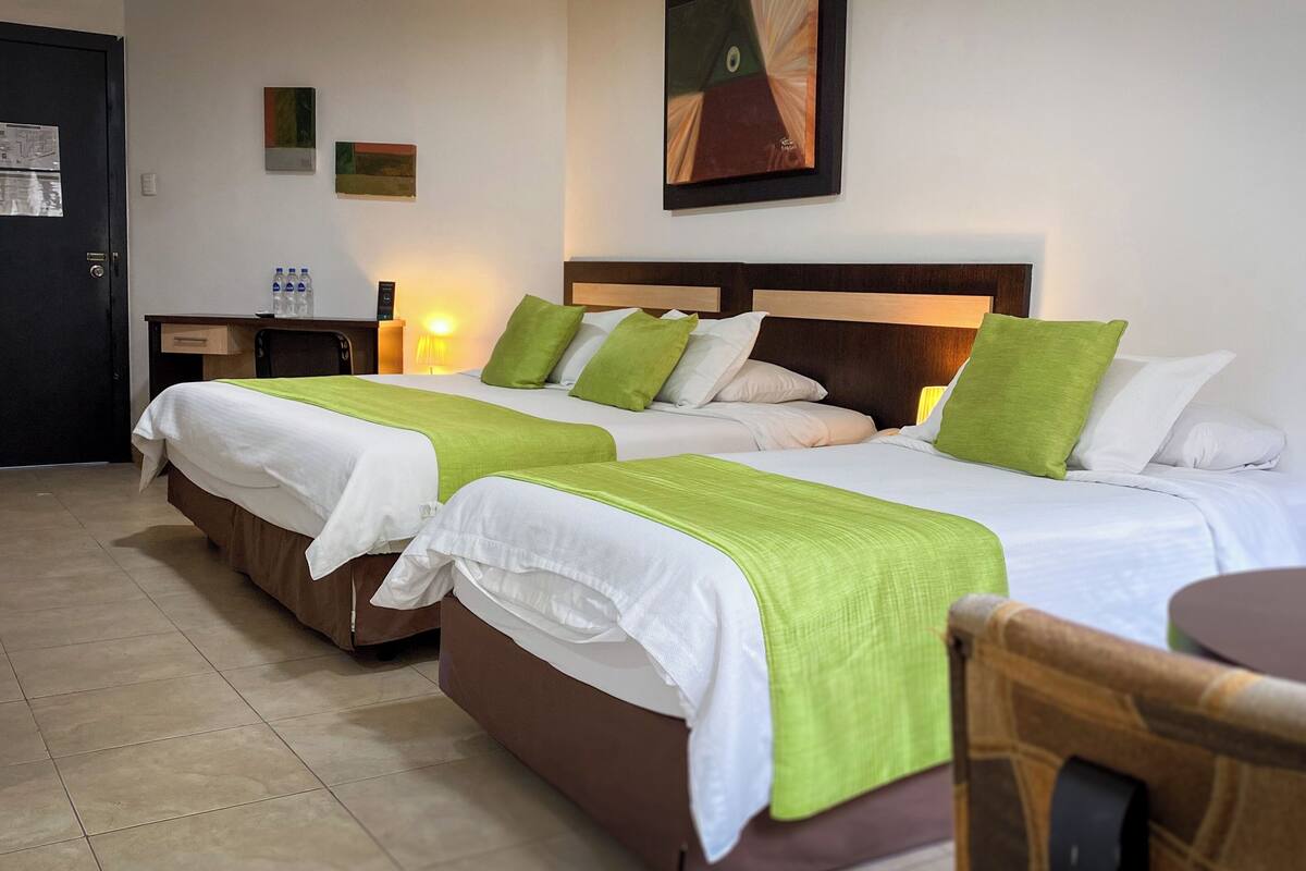 Habitación de hotel con dos camas, cada una con ropa de cama blanca y verde, un cabecero de madera y una pequeña mesa con una silla.