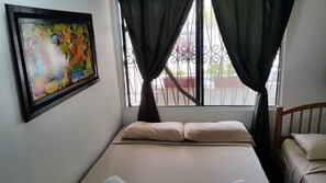 A bedroom with a bed, a window with curtains, a metal railing outside, and a colorful painting on the wall.