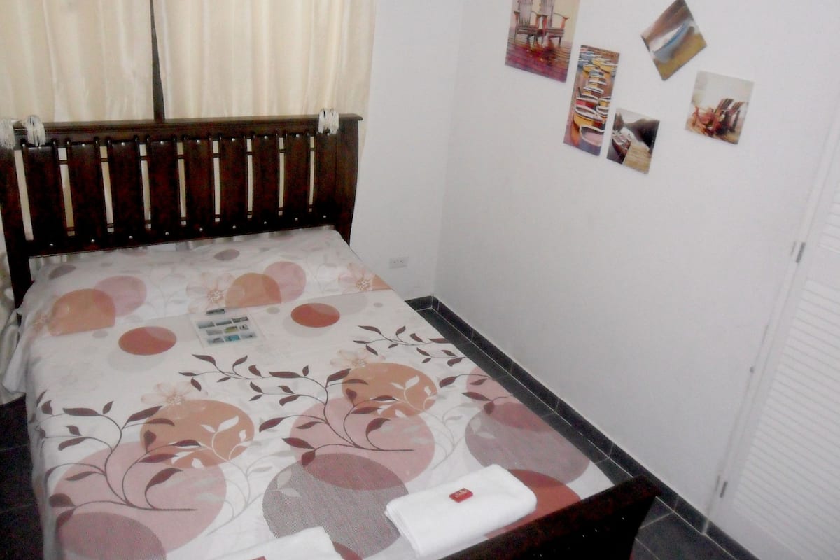 A bedroom with a wooden bed frame, a floral bedspread, and a wall with several small pictures.